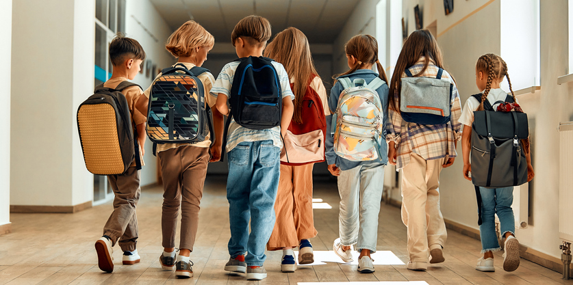 A group of children standing in a line, facing away from the camera with backpacks